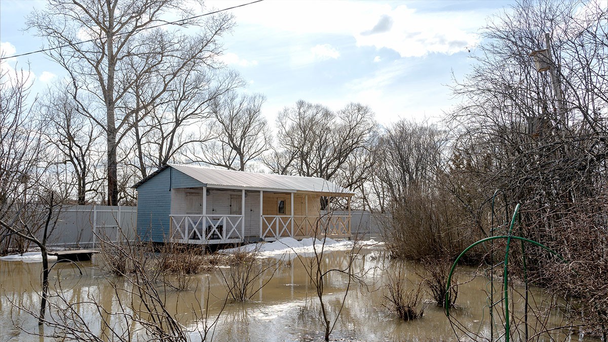 A small blue house on stilts surrounded by floodwater, bare trees, and a cloudy sky. Snow patches are visible on the ground.