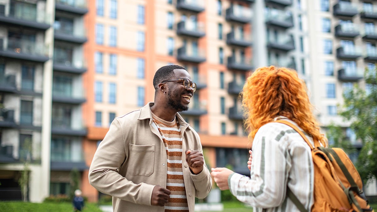 Two people smiling and talking in front of modern apartment buildings. One has a striped shirt, and the other has a backpack.