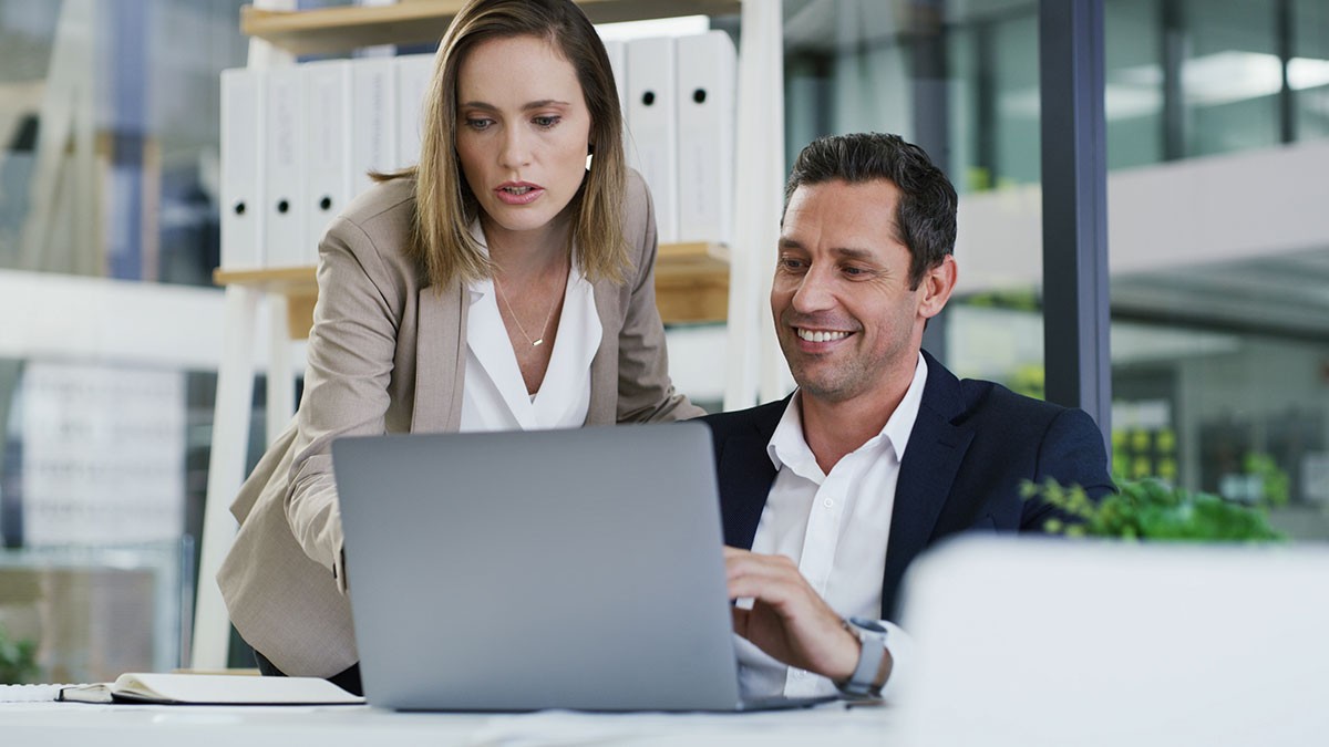 Two colleagues collaborate at a desk, with one pointing at a laptop screen and the other smiling, in a modern office setting.