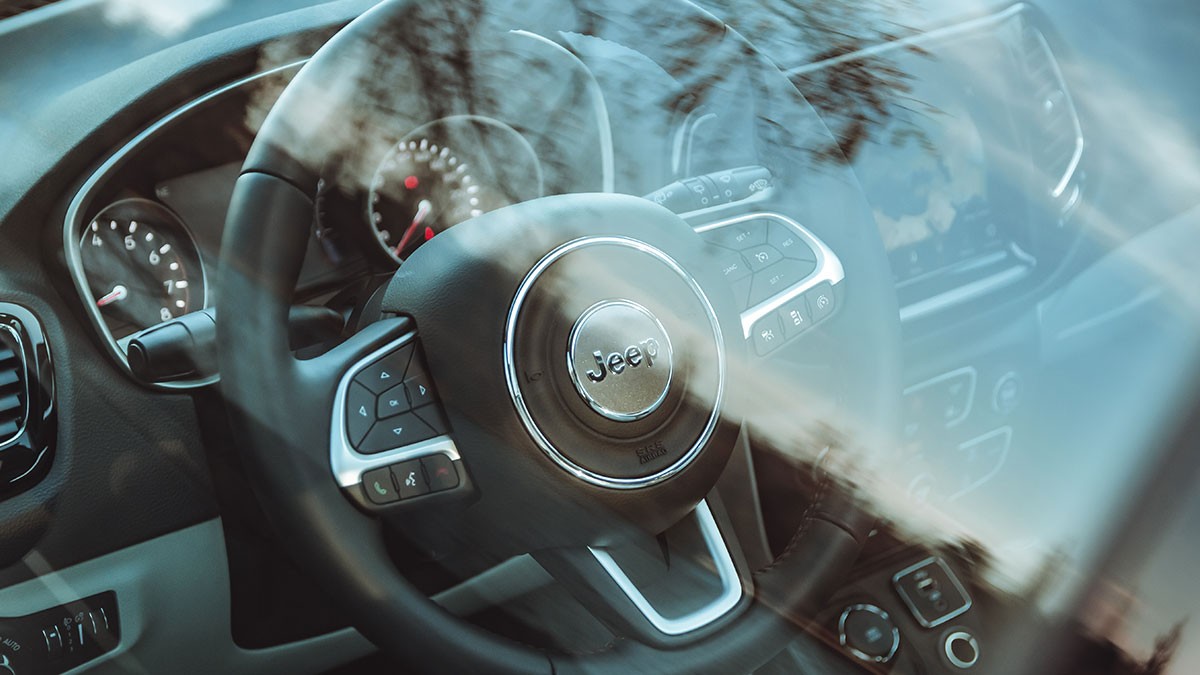 Close-up of a car interior showing a Jeep steering wheel, dashboard controls, and reflections of trees on the windshield.