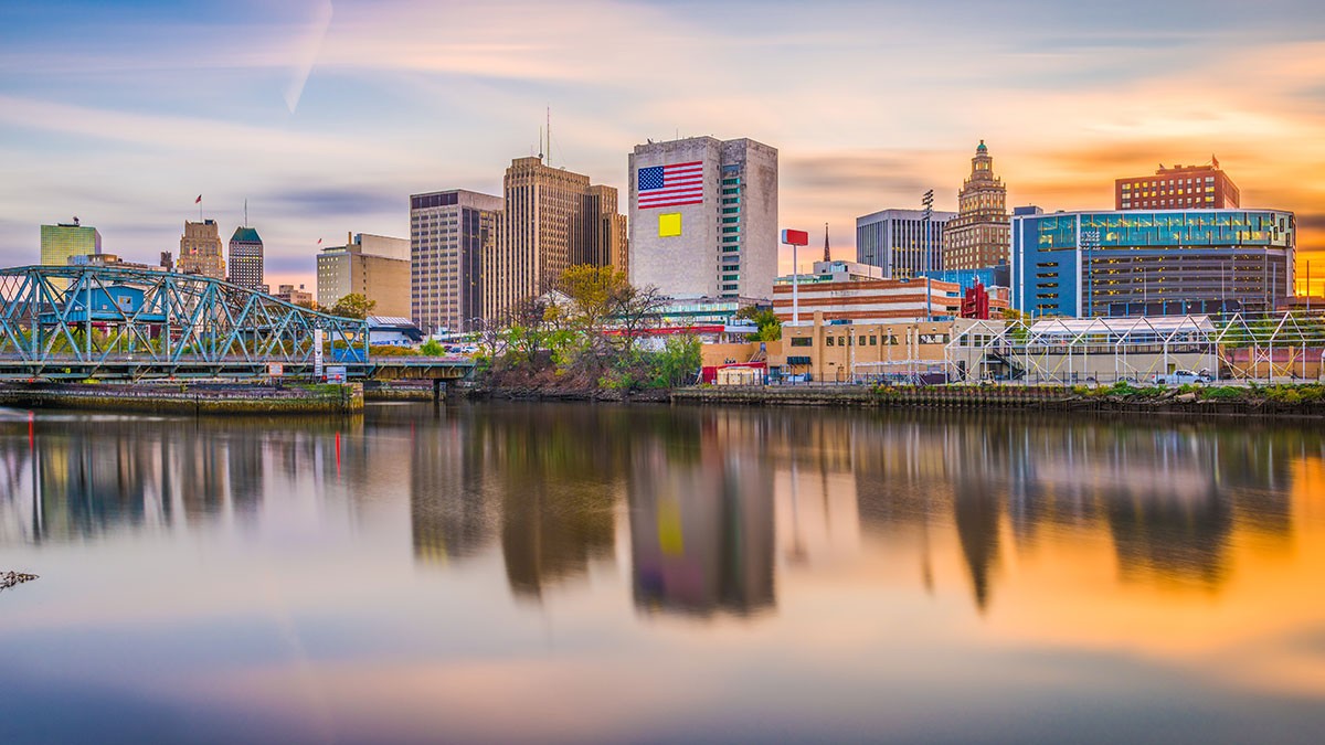 City skyline reflected in a calm river at sunset, with colorful clouds, tall buildings, and a bridge visible in the foreground.