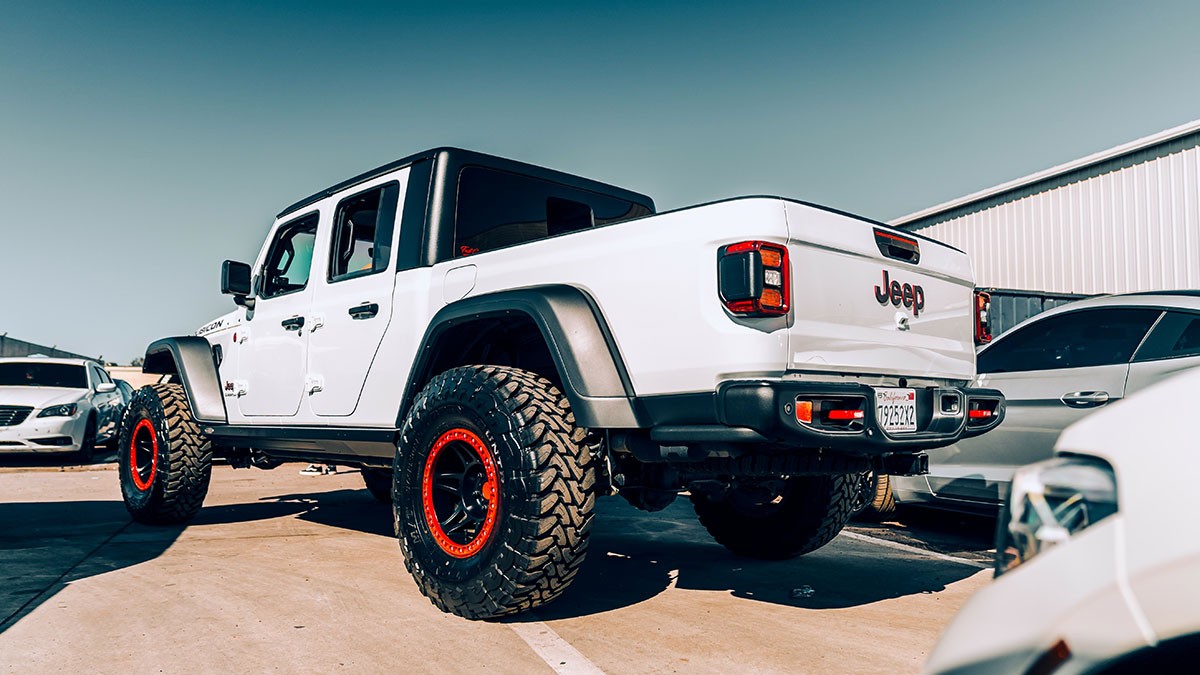White Jeep truck with large tires and red rims parked in a lot, viewed from the rear, under a clear blue sky.