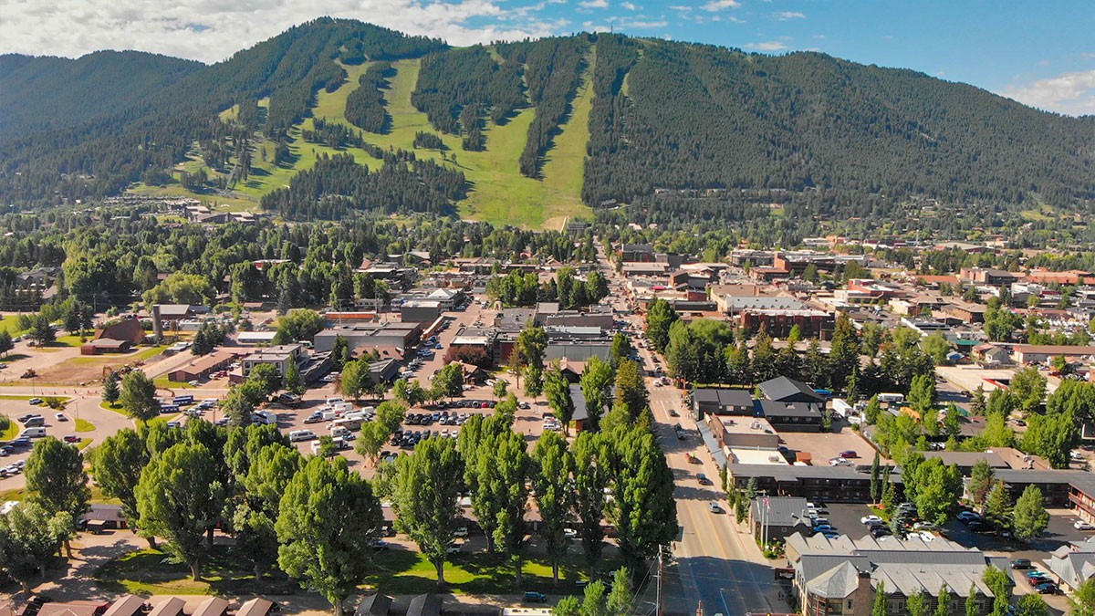 Aerial view of a town nestled near a mountain with ski slopes, surrounded by lush greenery and trees under a partly cloudy sky.