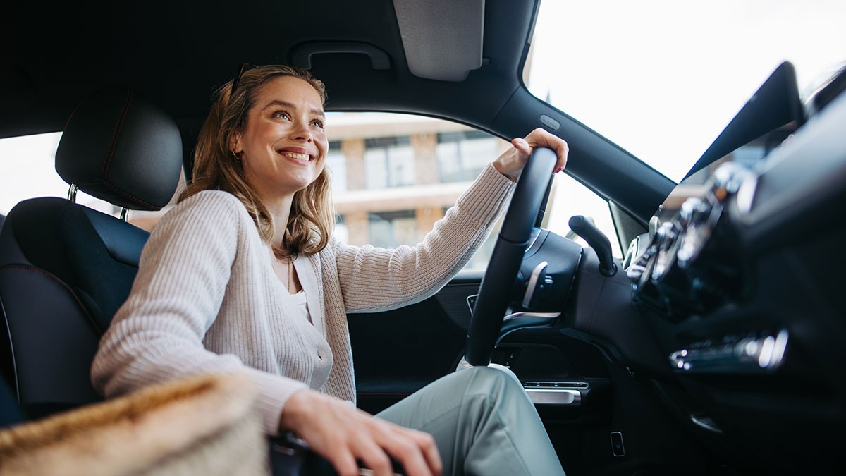 Smiling woman seated in a car, looking out the window, gripping the steering wheel, wearing a light sweater and surrounded by bright natural light.