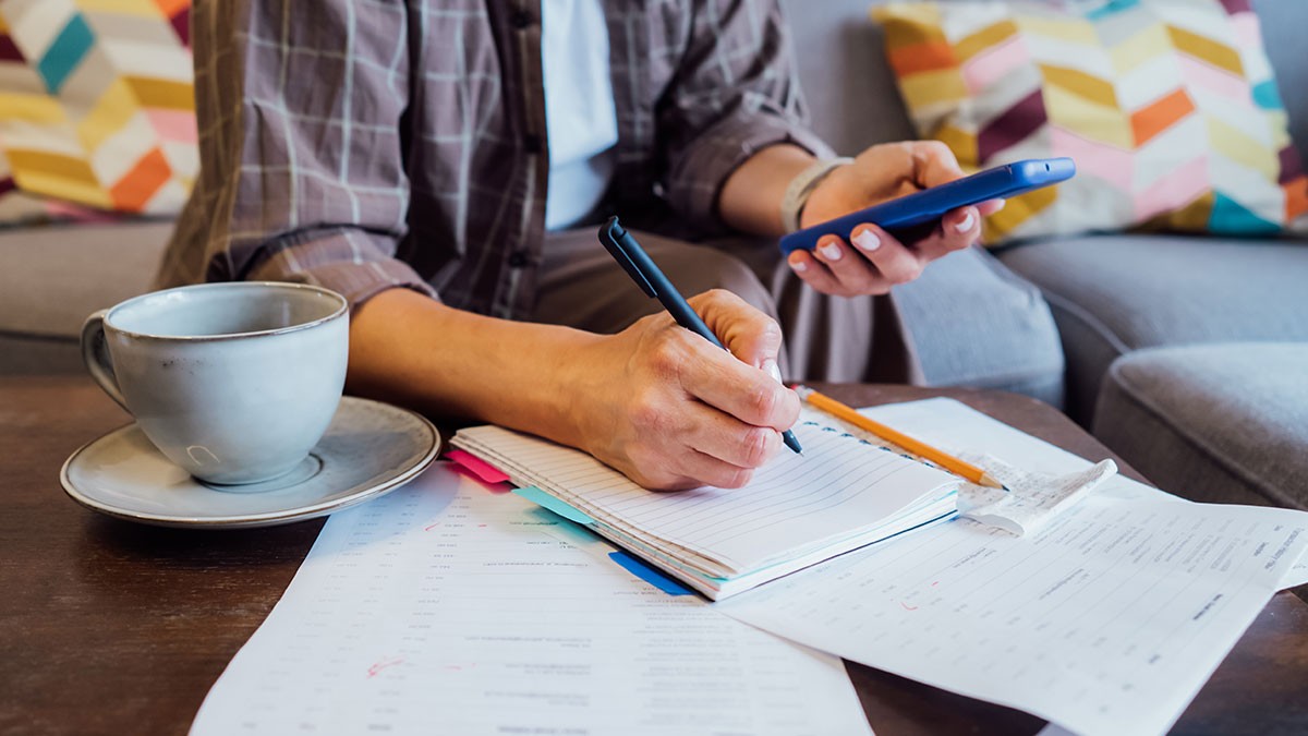 Person writing in a notebook while holding a smartphone, surrounded by papers and a coffee cup, seated on a couch with colorful pillows.