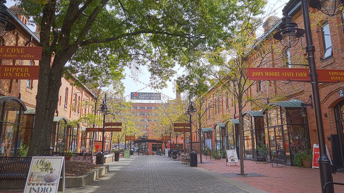 Brick-paved pedestrian street lined with red-brick shops, awnings, hanging signs, lampposts and leafy trees.
