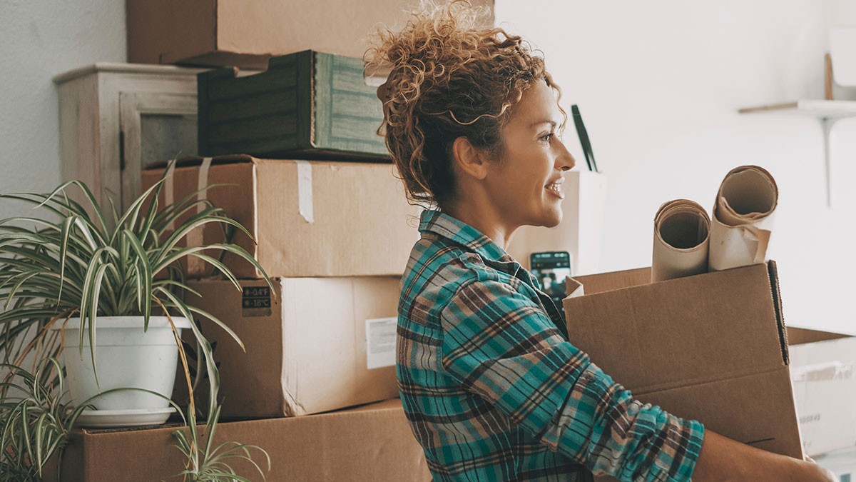 Woman smiling, holding a cardboard box with rolled papers, standing near stacked boxes and a plant in a room.