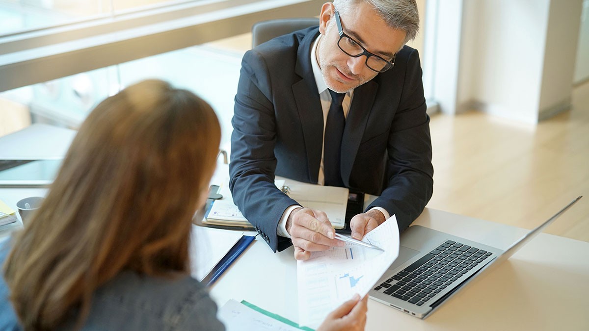 Two professionals in an office review financial documents, with a laptop open nearby.