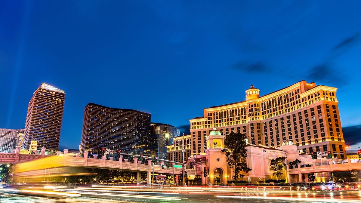Las Vegas skyline at dusk with illuminated hotels, including a large, brightly lit building, and light streaks from passing traffic in the foreground.