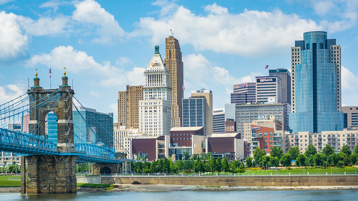 Skyline of Cincinnati with tall buildings, a blue suspension bridge, and a river in the foreground under a partly cloudy sky.