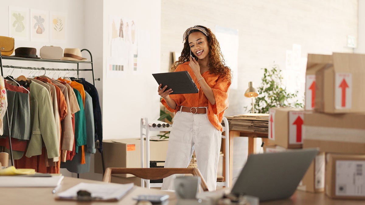 Woman in a bright office, holding a tablet, surrounded by clothing racks and boxes, smiling and wearing headphones.