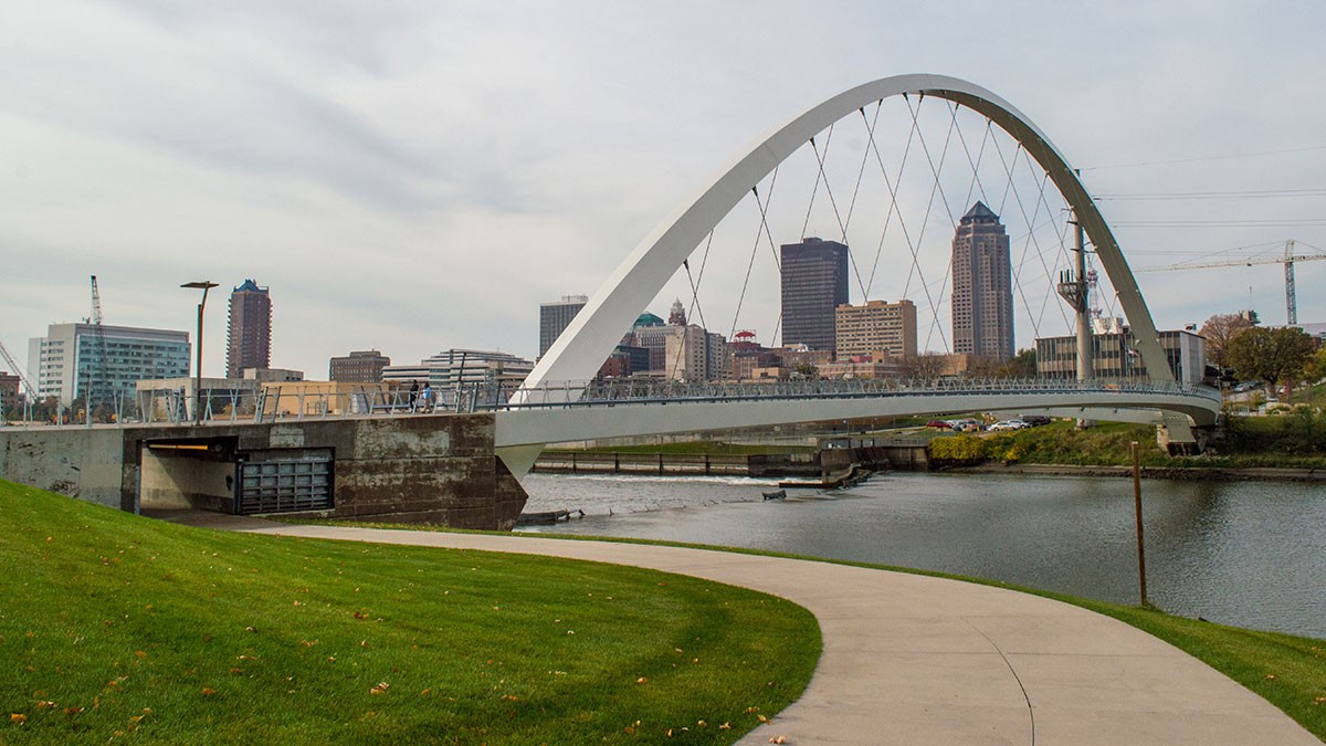 A modern arch bridge spans a calm river, with a city skyline in the background under a cloudy sky. A curved path leads to the bridge.