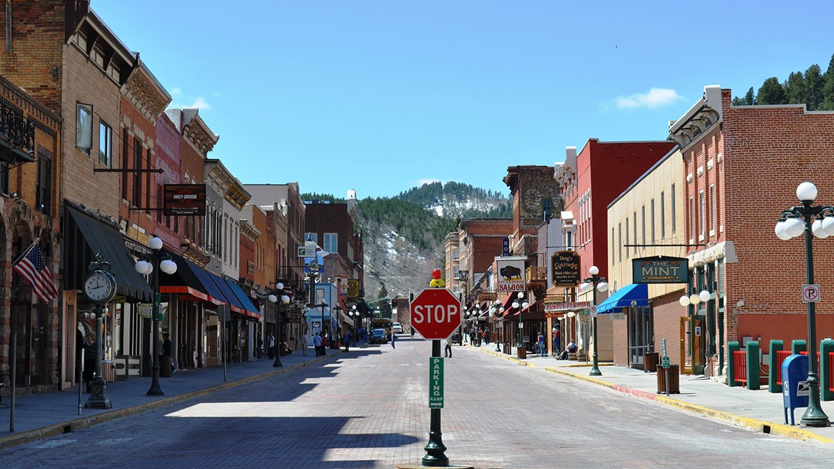 A quiet, empty street in a small town with historic brick buildings, a stop sign in the foreground, and snow-capped hills in the background.