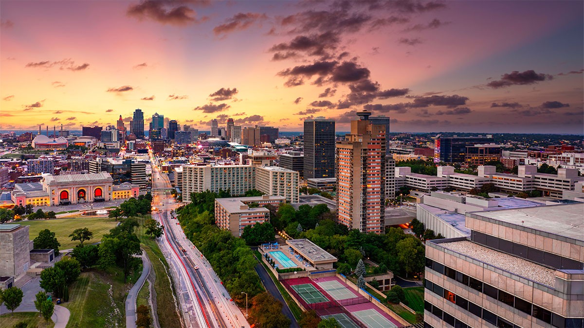 Aerial view of a cityscape at sunset, featuring tall buildings, roads, and tennis courts under a colorful sky with scattered clouds.