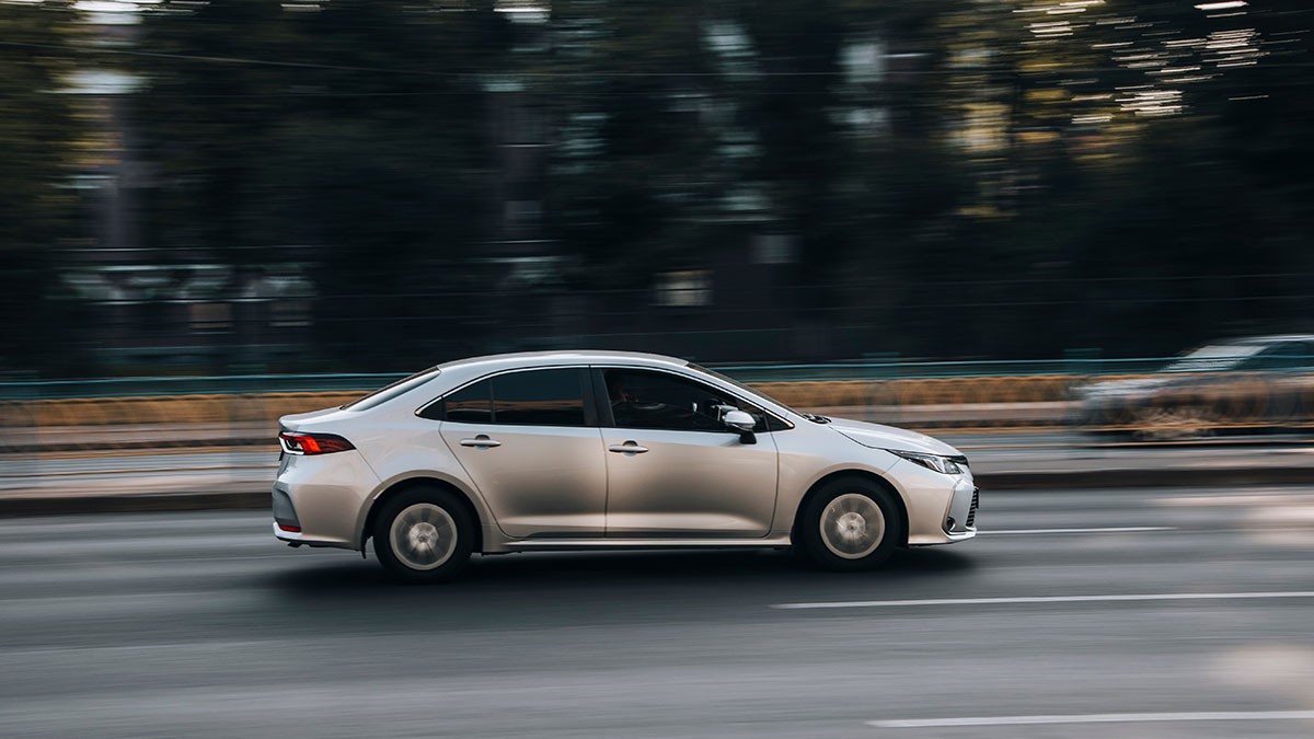 Silver sedan driving on a city street, motion blurred background with trees and buildings.