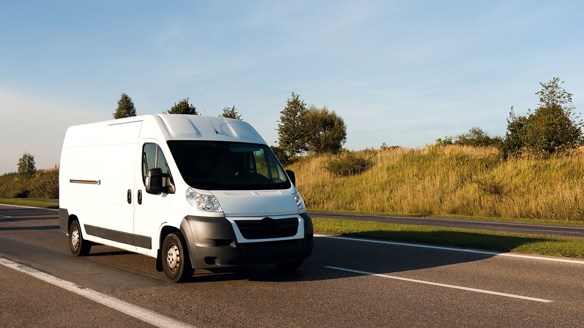 White delivery van driving on a rural road, surrounded by grass and trees under a clear blue sky.