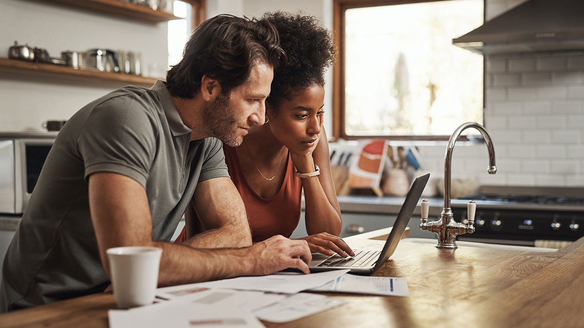 A couple leans over a kitchen counter, intently looking at a laptop. Papers are scattered around and a coffee mug sits nearby.