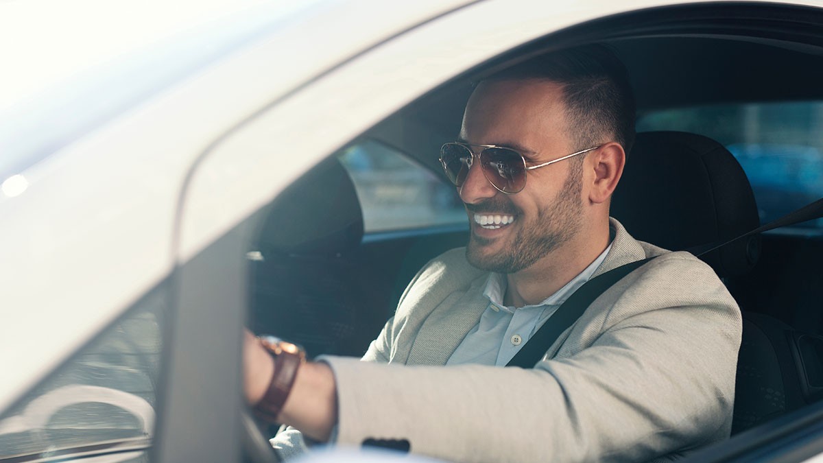 Man wearing sunglasses and a beige jacket smiles while driving a car, with sunlight streaming through the window.