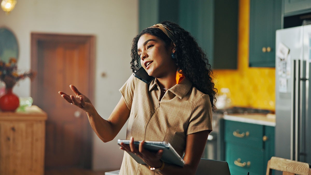 Woman multitasking in a kitchen, talking on the phone and holding a tablet, with a relaxed expression.