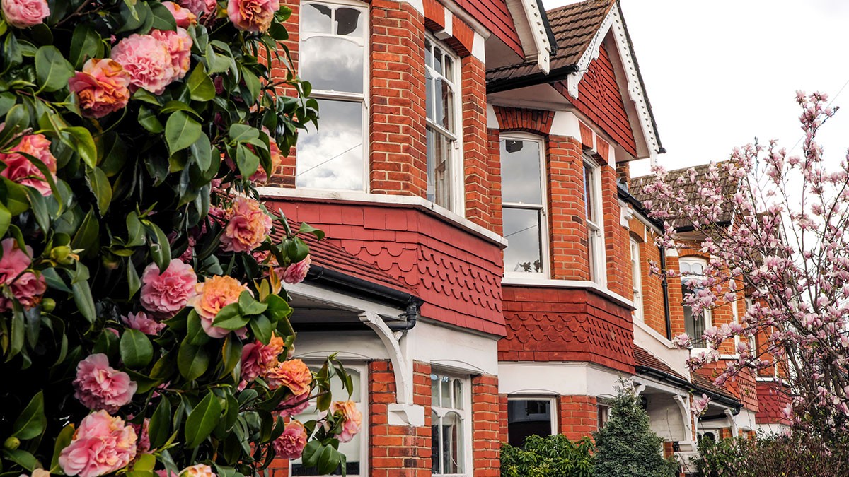 Row of charming red brick houses with white trim, surrounded by blooming pink flowers and lush greenery under a cloudy sky.