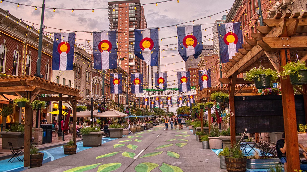 A vibrant street scene with colorful flags, outdoor seating, decorative lights, and painted pathways, surrounded by brick buildings.