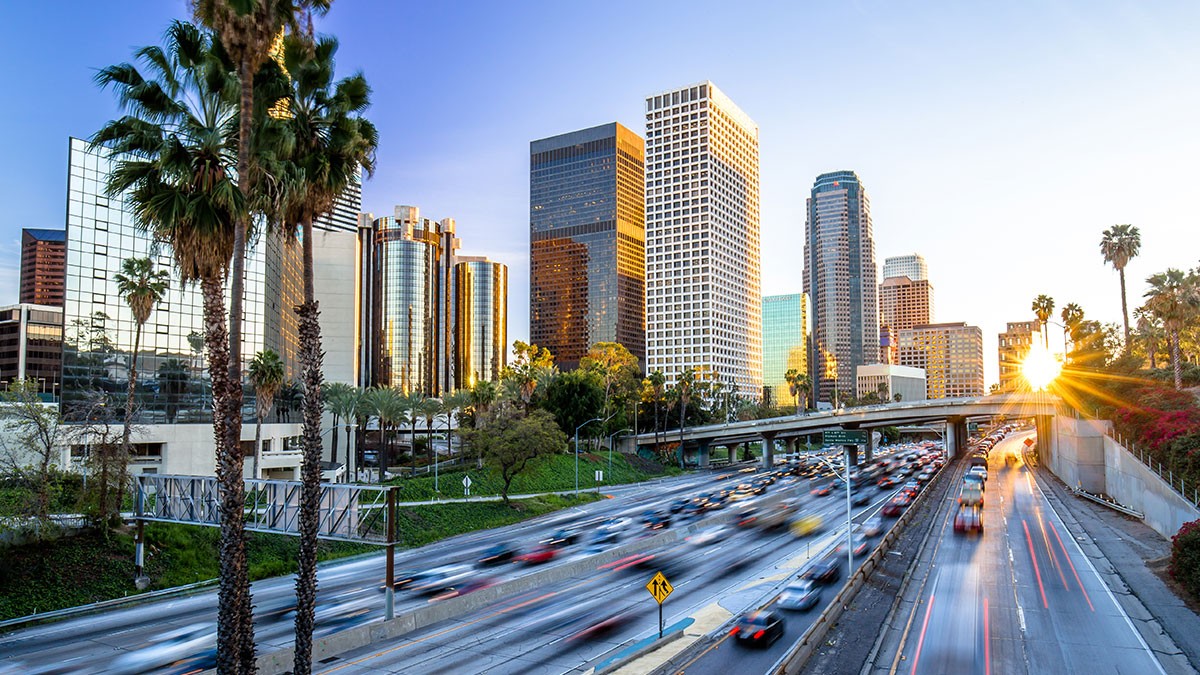 Sunset view of a busy highway with blurred traffic, surrounded by tall skyscrapers and palm trees in a cityscape.