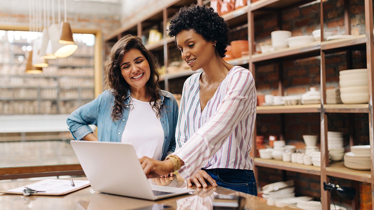 Two women smiling and looking at a laptop in a pottery shop, surrounded by shelves of ceramic items.