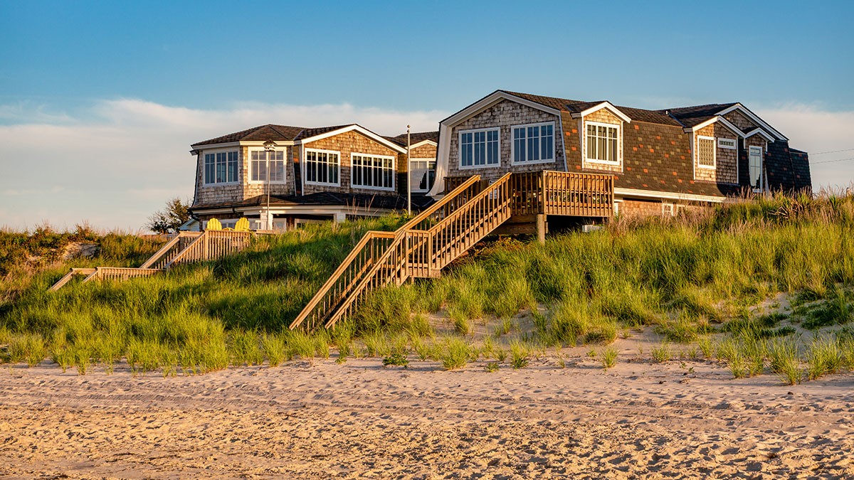 Shingled beach house with large windows and wooden stairs, surrounded by green grass, overlooking a sandy beach under a clear blue sky.