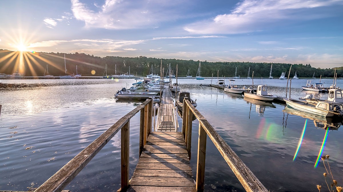 A wooden dock extends into a calm lake with boats moored on either side. The sun sets on the horizon, casting a warm glow over the scene.