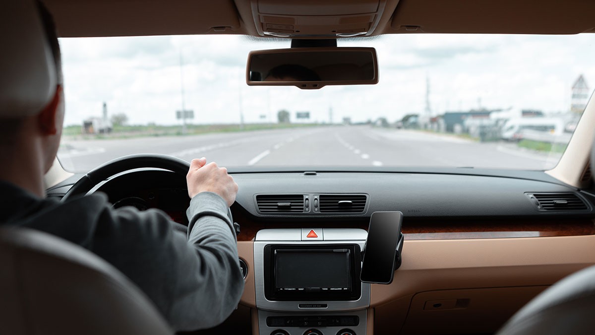 Driver in a car on a clear, open highway, viewed from the back seat, with hands on the steering wheel and a cloudy sky outside.