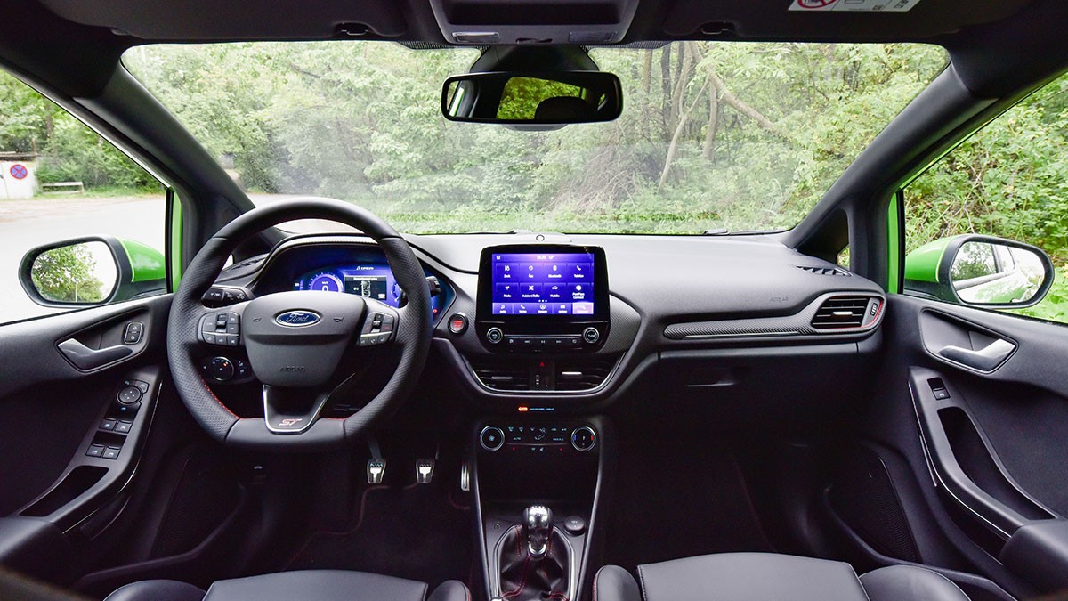Interior view of a car with a green exterior, featuring a modern dashboard, touchscreen display, and black steering wheel, set against a forest backdrop.