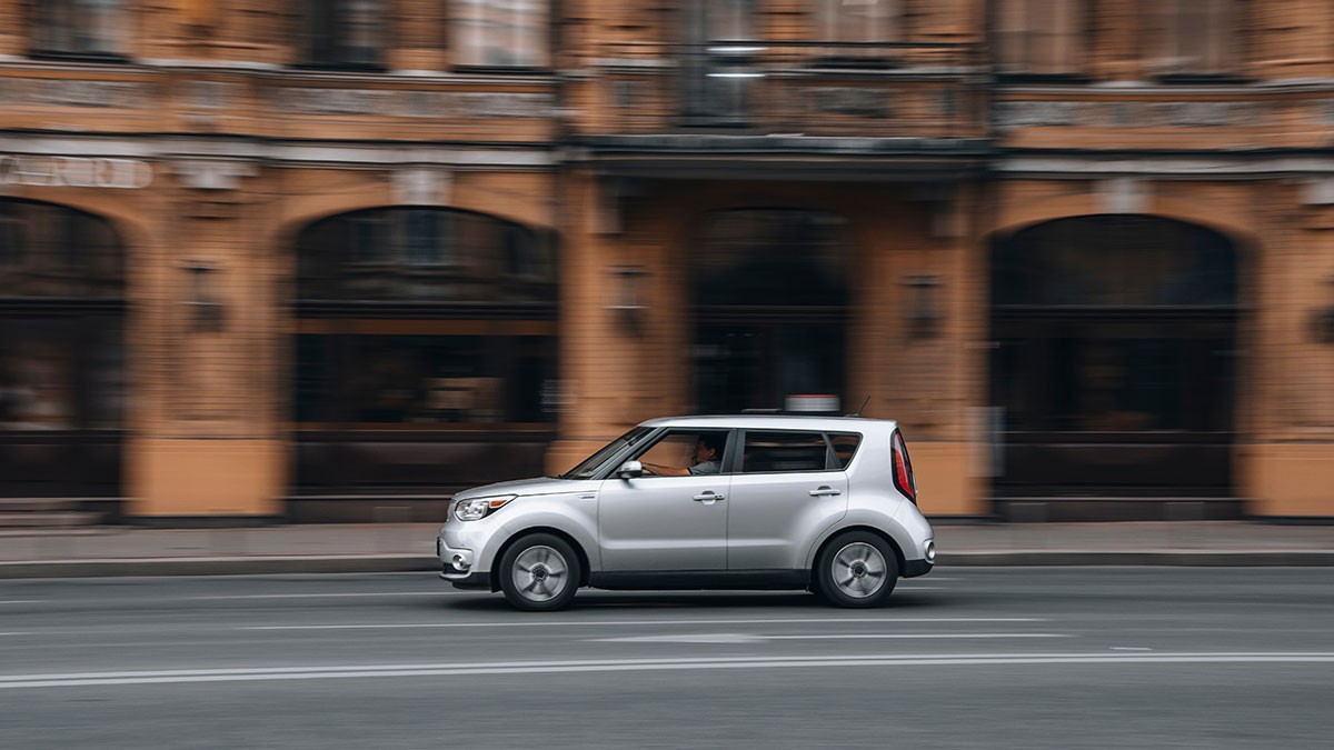 Silver compact car driving past a blurred historic building with arched windows on a city street.