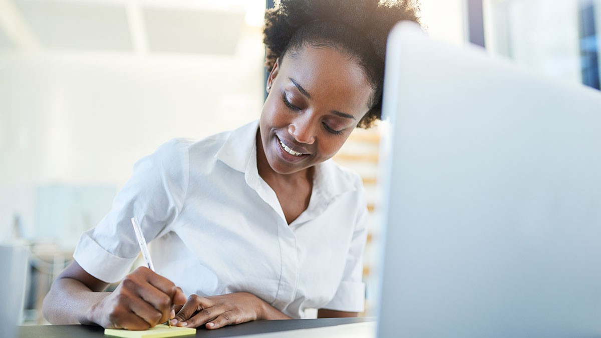 Woman in a white shirt writing on a notepad at a desk, with a laptop in the foreground. Bright, well-lit office environment.