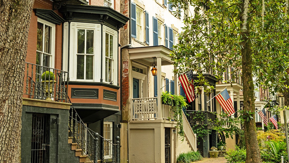 Historic row houses with bay windows and American flags, lined with trees and lush greenery on a sunny street.