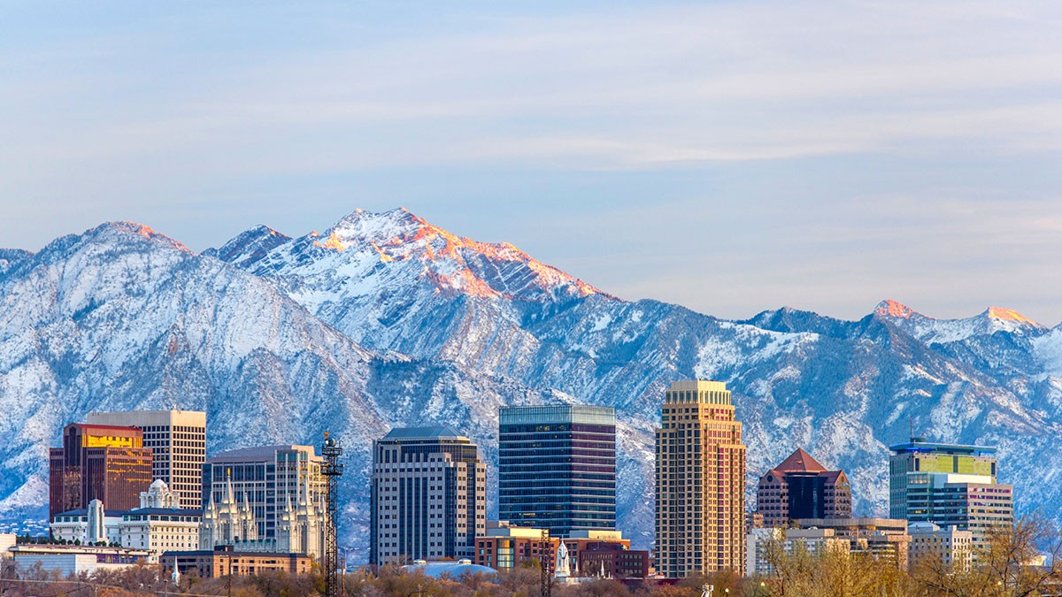 City skyline with tall buildings, set against a backdrop of snow-covered mountains under a partly cloudy sky.
