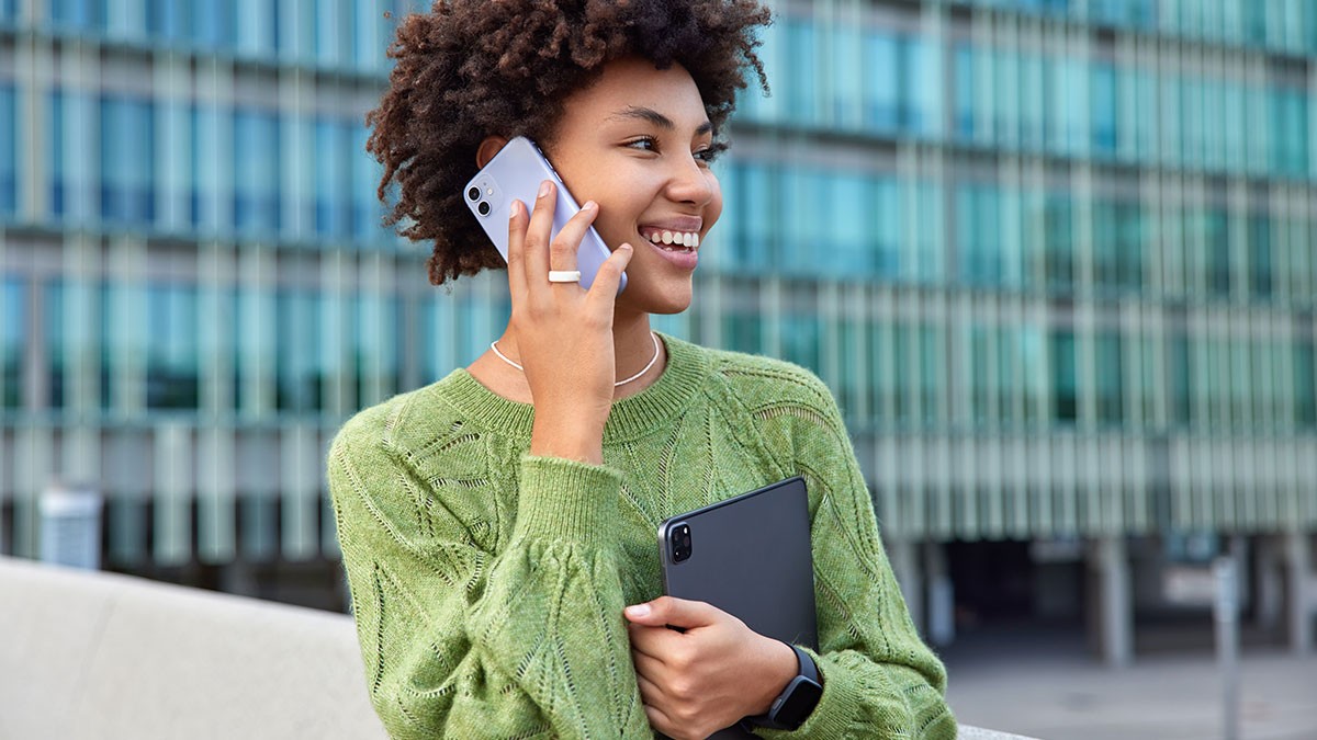 Smiling person with curly hair in a green sweater holds a phone to their ear and a tablet, standing outside a modern glass building.