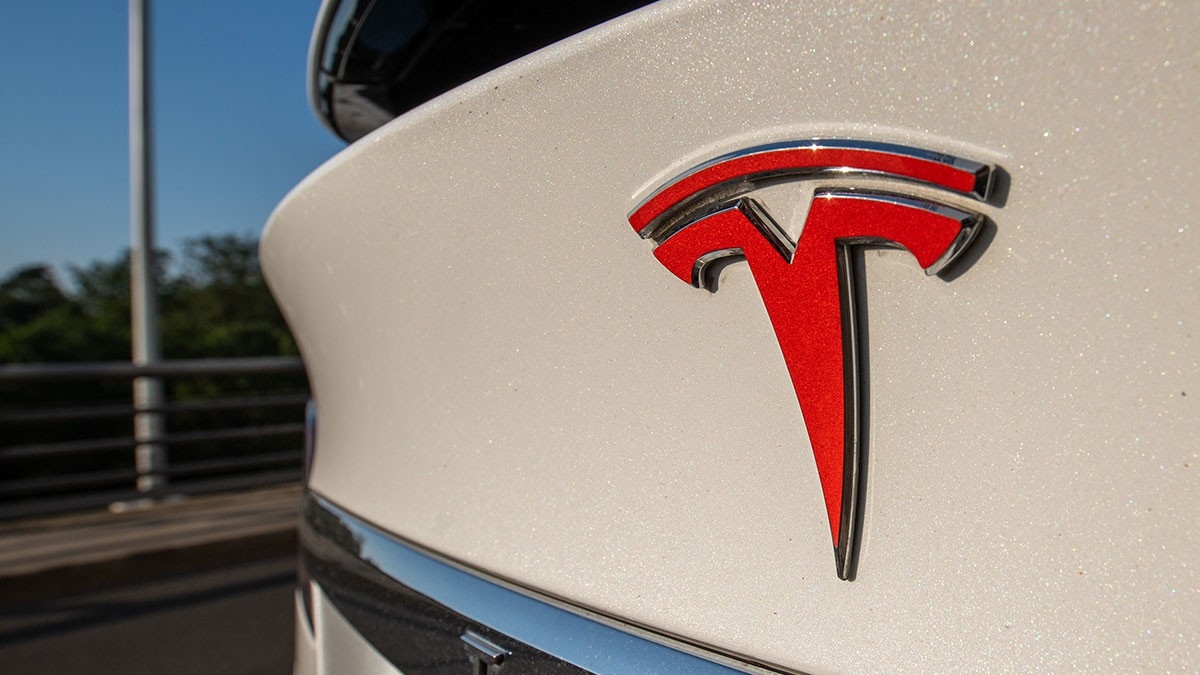Close-up of a red Tesla logo on a white car, with a blurred background featuring a railing and greenery.