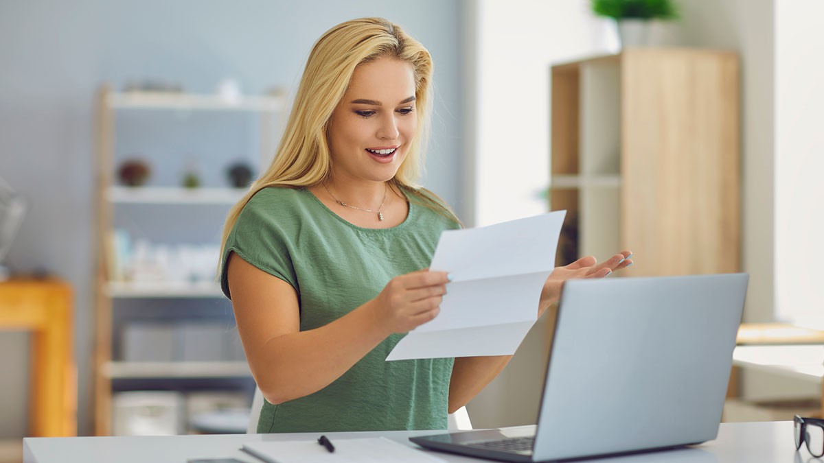 A woman in a green shirt smiles while reading a letter at her desk, with a laptop open, in a bright, modern office space.