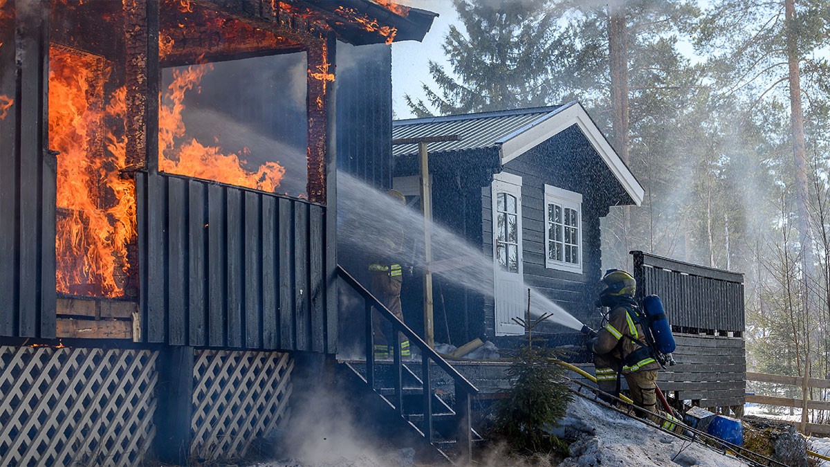 Firefighters extinguishing a blazing fire on a wooden house, surrounded by smoke and trees in the background.