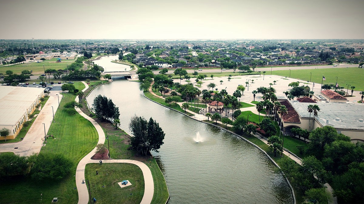 Aerial view of a park with a winding river, fountain, pathways, trees, and surrounding buildings under a cloudy sky.