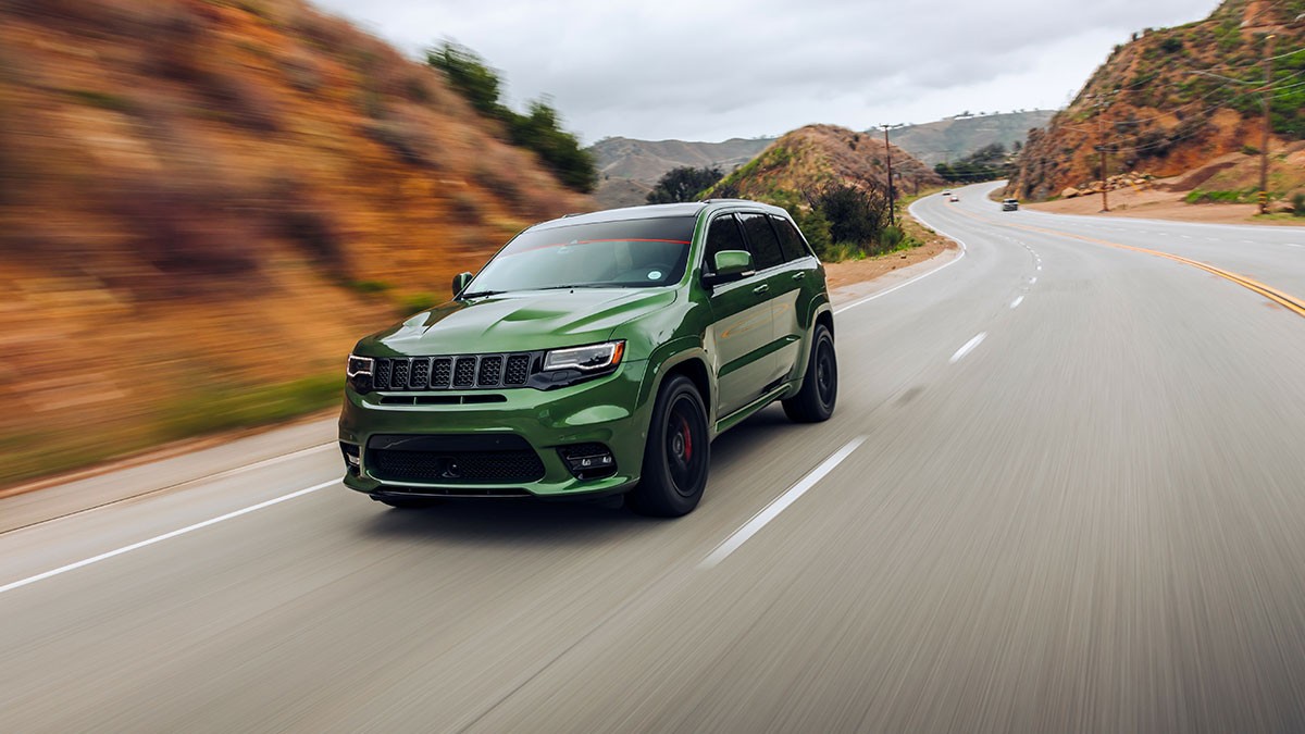 A green SUV drives on a winding mountain road, surrounded by rocky hills and overcast skies.
