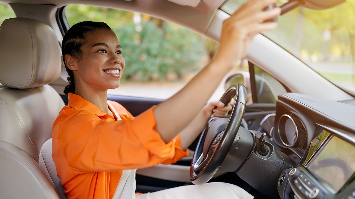 A person in an orange shirt smiles while adjusting the rearview mirror inside a car, with trees visible through the windows.