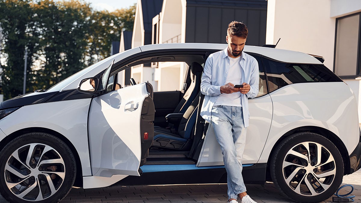 Man leaning on a white car with an open door, looking at his phone, outdoors on a sunny day.