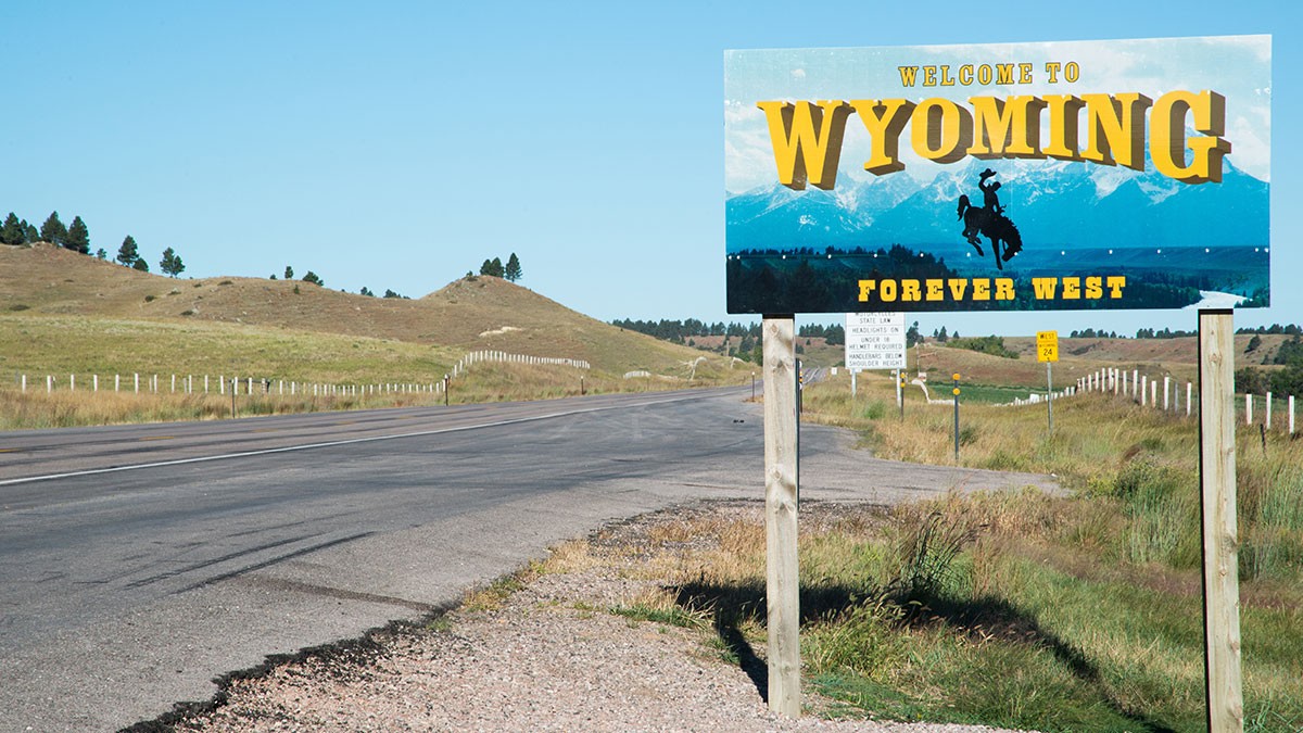 Roadside sign reading "Welcome to Wyoming, Forever West" with a cowboy silhouette, against a backdrop of open fields and clear blue sky.