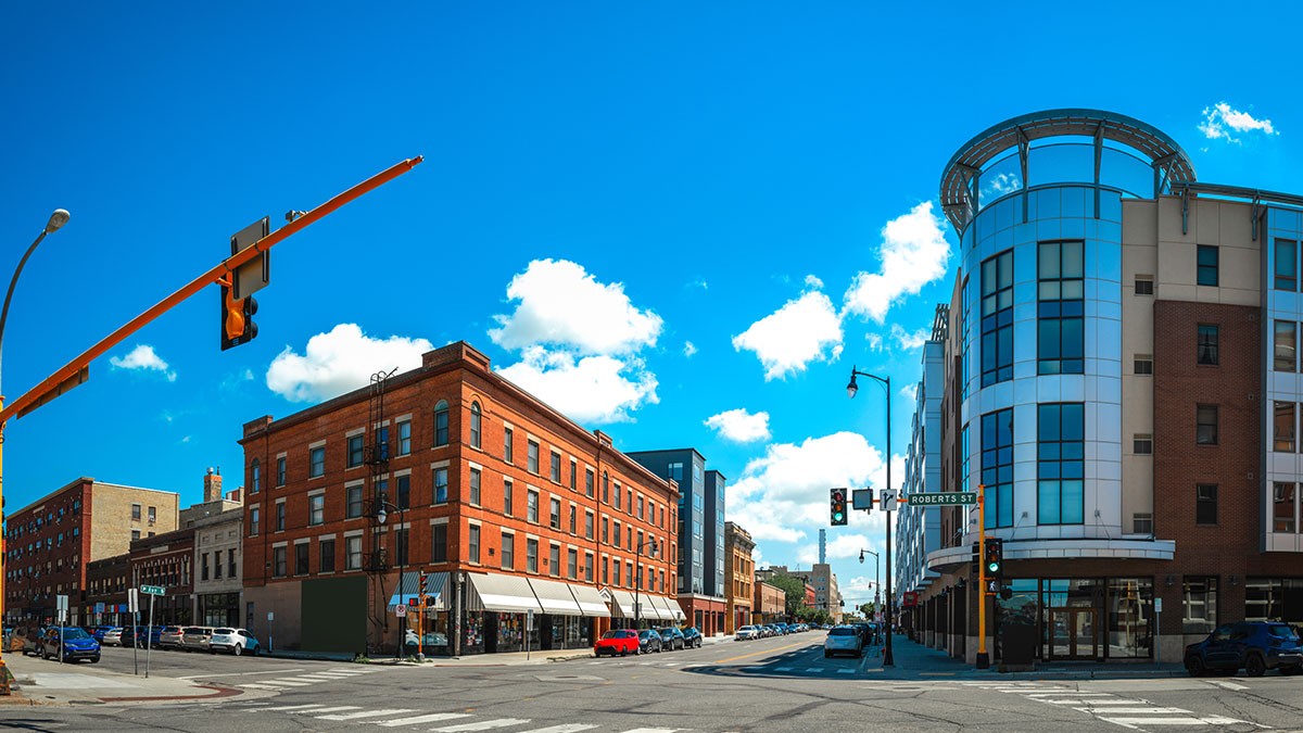 Street intersection with brick and modern buildings under a clear blue sky. Traffic lights and crosswalks are visible.