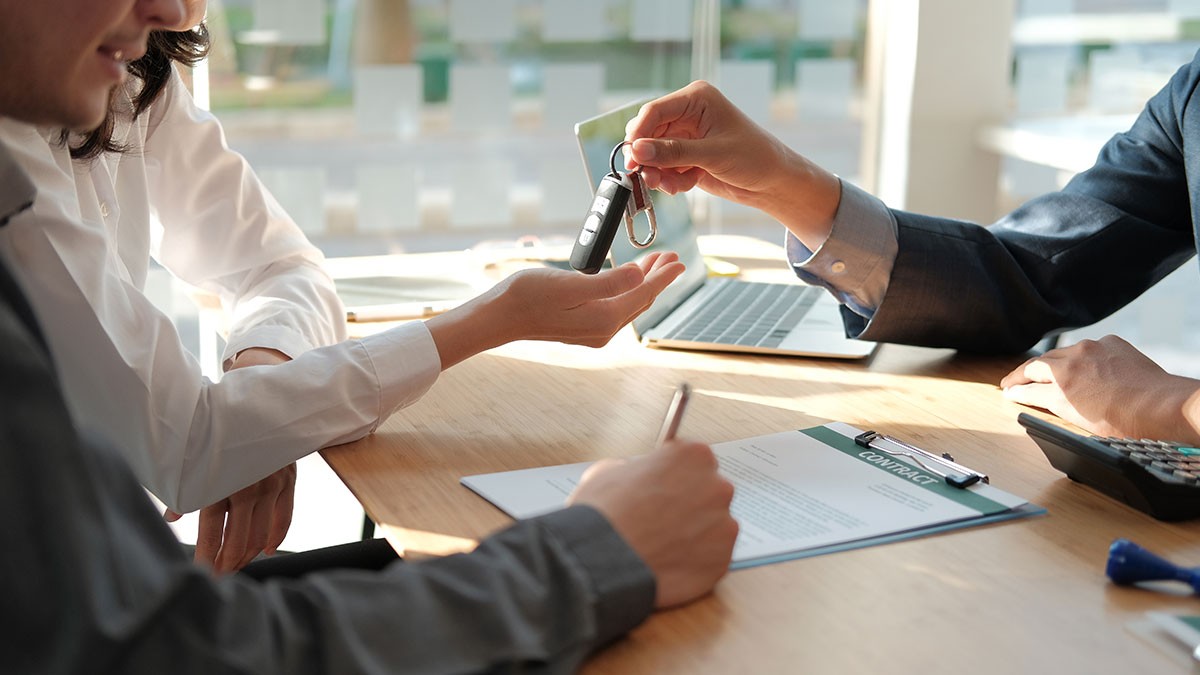 Person signing a document while receiving car keys from another individual in an office setting, signifying a transaction or agreement.