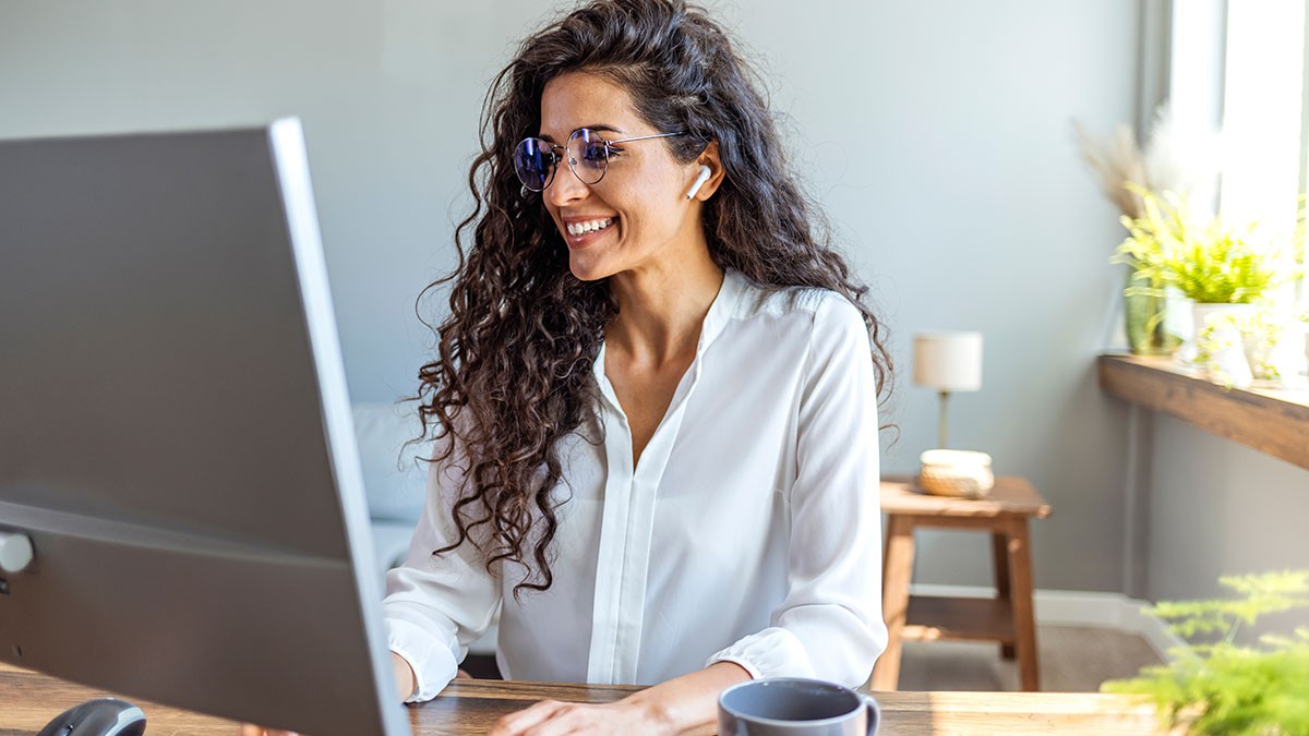 A woman with long curly hair, wearing glasses and earbuds, smiles while working at a computer in a bright home office with plants.