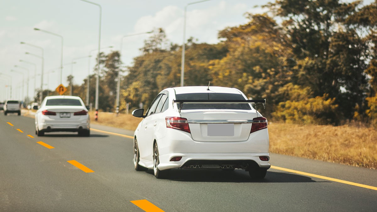 White car with a rear spoiler driving on a highway, surrounded by trees and other vehicles under a clear sky.