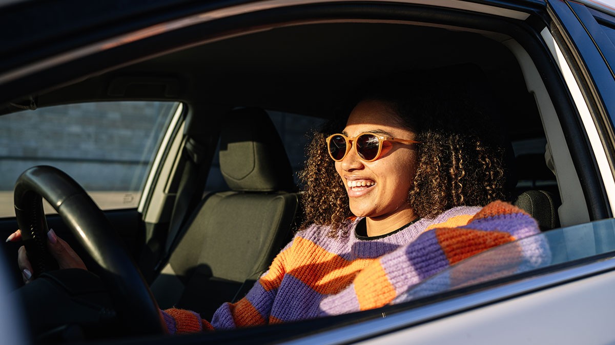 Person with curly hair and sunglasses smiling while sitting in a car, wearing a colorful striped sweater, with sunlight illuminating their face.