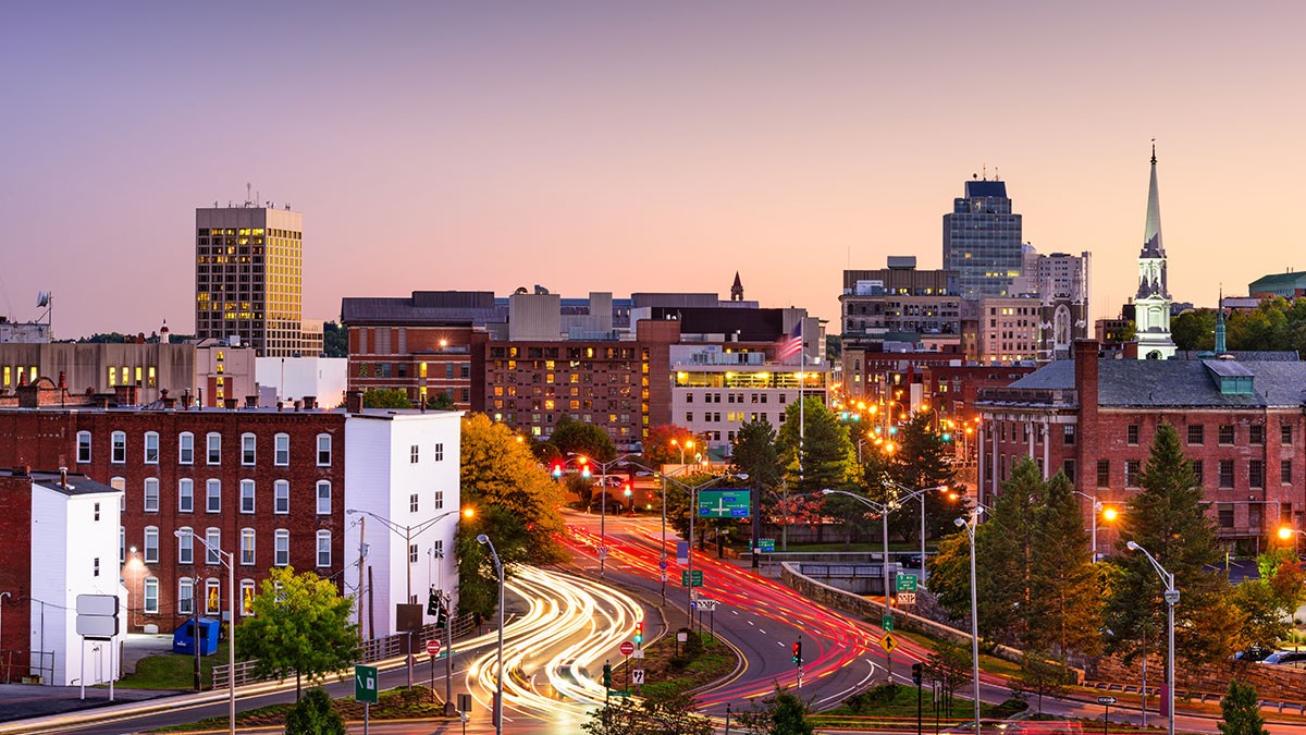 Cityscape at dusk with light trails from traffic, buildings, and a church steeple against a purple and orange sky.